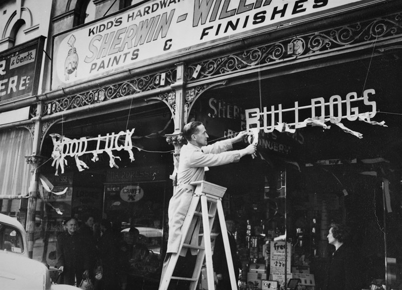 A black and white photograph of a man up a ladder outside a shop.