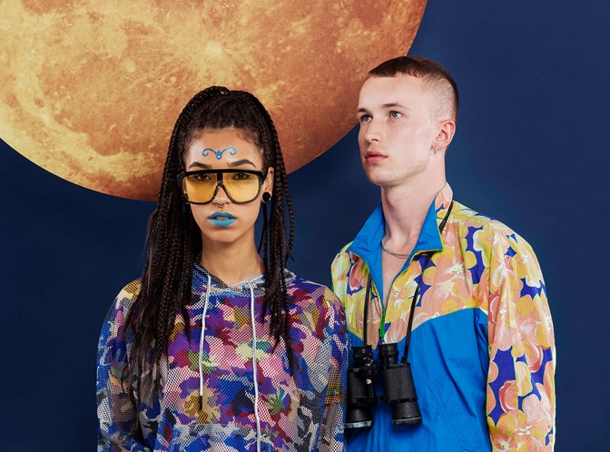 A young woman and man, each dressed in vivid colours, standing in front of a picture of a celestial body. She holds a thermos; he, binoculars.