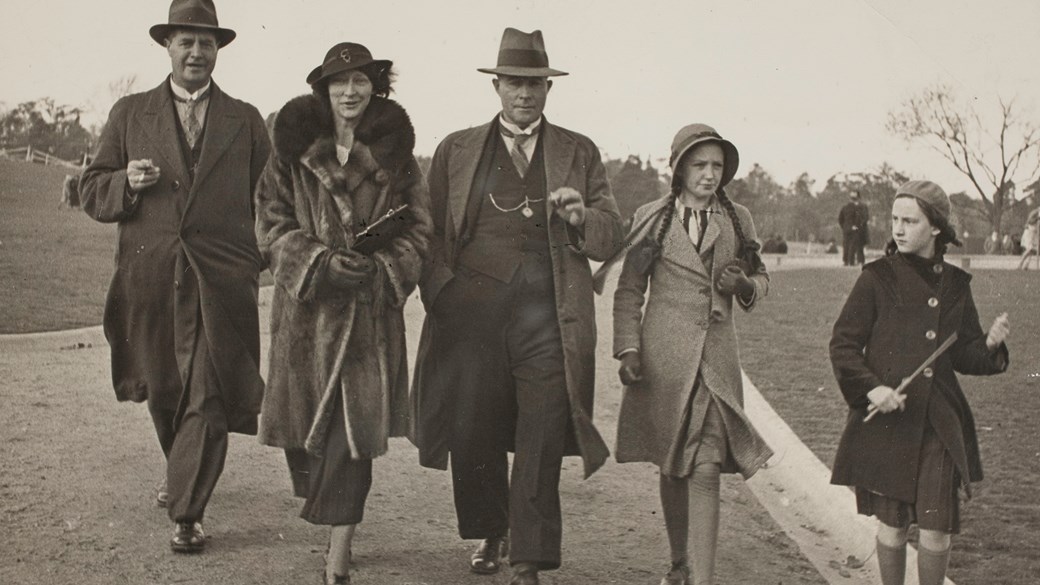 Two men, one woman and two girls in winter coats walking along a gravel path in a park. All are wearing hats.