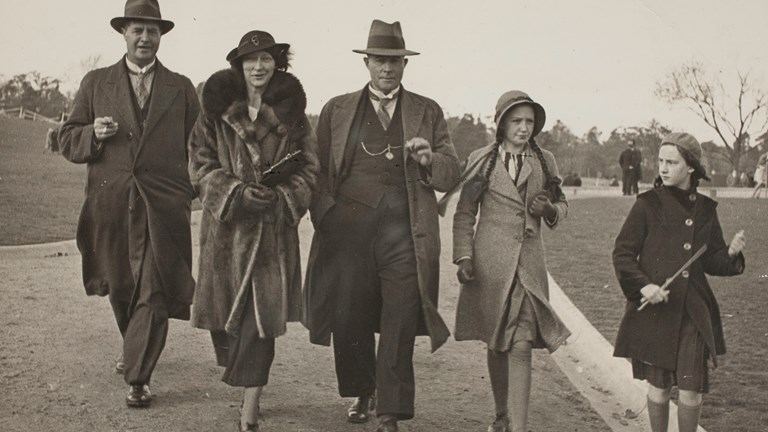 Two men, one woman and two girls in winter coats walking along a gravel path in a park. All are wearing hats.