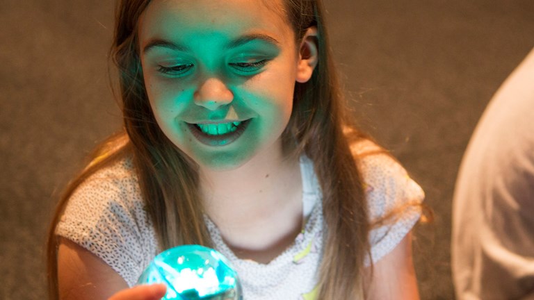 A child holds a Sphero® robot in her hand. The robot is the size and shape of a tennis ball. There is a light shining from inside.