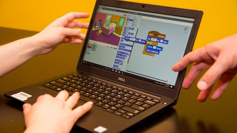 A laptop computer screen shows a game being programmed. An adult’s and a child’s hands are pointing at the screen.