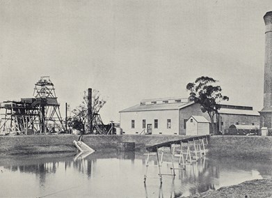 Water reservoir with poppet head, mine buildings and tall chimney in background.