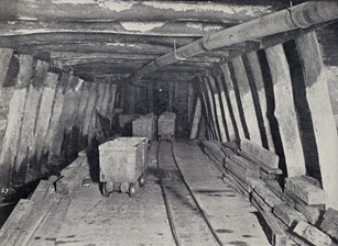 Underground chamber in mine shaft with small carts on a rail line, and a pipe overhead.
