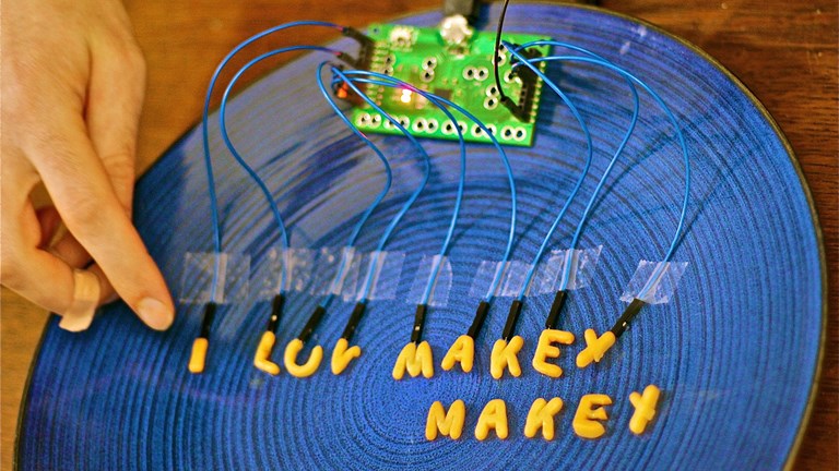 A person’s finger is about to touch a piece of dry alphabet soup. The alphabet soup pieces are connected by wires to a Makey Makey electronic board. The letters spell out “I luv makey makey
