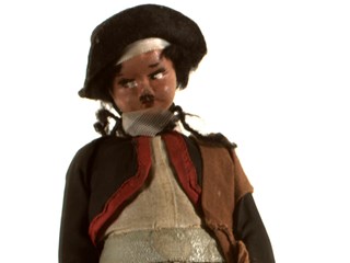 Doll dressed in a grey hat, black boots, black and white checkered pants, a white shirt and a black bolero jacket. He also has a piece of brown fabric draped over his left shoulder, a piece of leather around his right wrist and a belt with three wooden balls is around the doll's waist. The doll has dark hair.