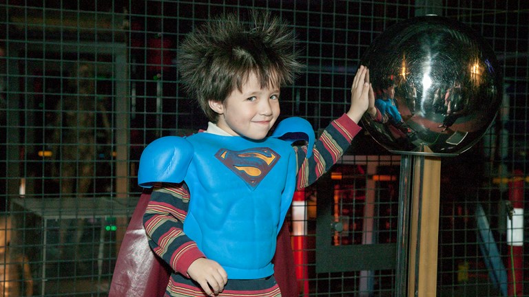 Boy wearing a Superman costume standing on the Van de Graaff generator in the Lightning Room at Scienceworks
