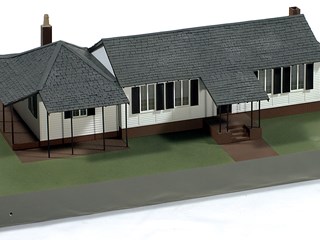 Built on a sloping base, the model depicts a white-painted weatherboard cottage with two red brick chimneys and red brick foundations.