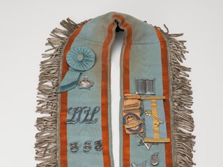 Collar - pale blue, ribbed satin with embossed orange trim. There are several jewels and badges pinned to the collar. 