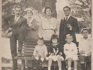 Four adults standing and four children on a park bench. There are hats and umbrellas in the grass in front of them.