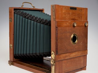 A wooden boxed bellows camera. The camera is made of wood and metal (brass) and would have once had a glass lens. There is a plaque with the makers details on the front of the camera.