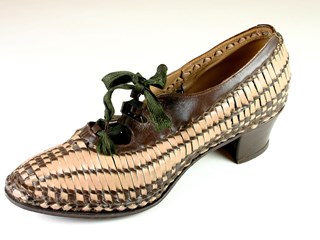 Left foot low heeled female dress shoe of pink and brown leather basketweave. Enclosed shoe with slight rectangular shaped toe, tied with green shoe lace. Lower shoe section and central section from base to vamp are woven in a checkered pattern, top portion appears as two rows of vertical stripes, and topline, including three band strapping area, is in unwoven brown leather