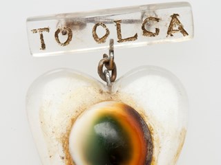 Brooch in shape of a heart with a round decoration. The words 'TO OLGA' are inscribed on a rectangular badge attached to the top of the brooch. There is a small loop of chain and a safety pin attached to the top of the brooch.