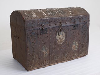 Rectangular shaped trunk made from wood and lined on the exterior with metal sheeting. The trunk has a domed lid, and features decorative stud work. The sides of the trunk also have stencilled images. The Inside of the trunk is lined with purple and white striped fabric. There is extensive text which has been painted onto the lid in white.