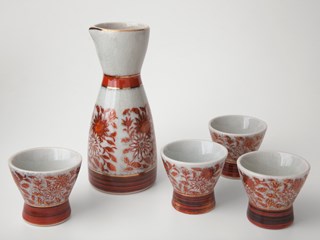 Ceramic sake carafe, off-white with orange floral design around the outside with an orange band at the neck and base of the carafe. The rim has a small pouring lip. Also four small sake cups in matching off-white and orange floral design.