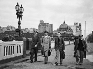 Four men walking on Princes Bridge, with Flinders Street Station and city buildings in the background. Gas lamps on the bridge are at left. A car is on St Kilda Road at right.