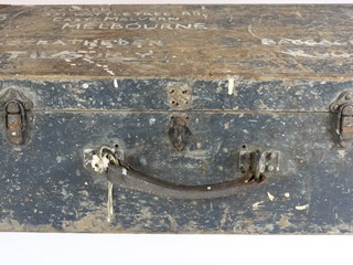 Small wooden truck. Writing in white paint on the lid and left side of trunk. Remnants of dark green paint on outside of trunk, grey inside. Large ring marks on lid. Blue, white, purple and black P & O baggage label on the right side of trunk. Brown leather handle. Steel corners, central hasp and staple (missing hasp) and two clasps. Four rubber feet on bottom.