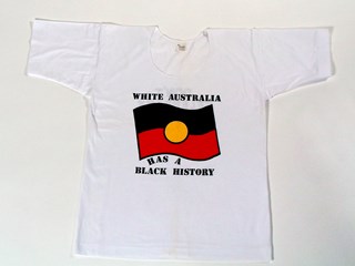 White cotton t-Shirt with a colour screen print of the Australian Aboriginal flag (rectangle with black top half, red lower half and yellow circle in the centre). The T-Shirt has black text on the back and front: "White Australia has a Black History"