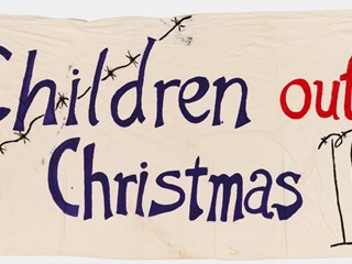 Banner of cream soft linen with purple, red and black painted slogan 'Children out for Christmas' with image of razor wire and 3 figures behind fence with razor wire. Both sides stapled sleeve for threading through carry poles.
