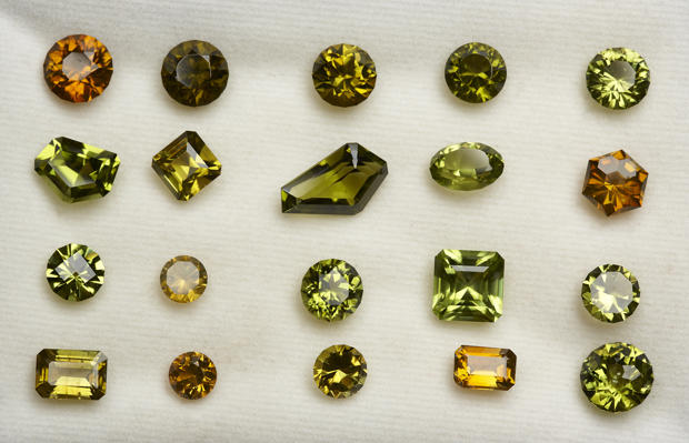 Twenty faceted forsterite specimens in a range of green and yellow hues.