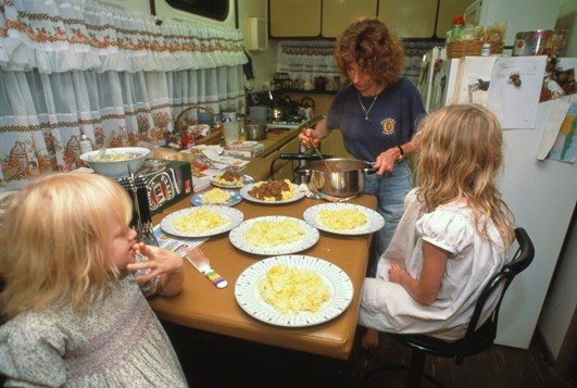 Woman serving food from a saucepan as two small children watch