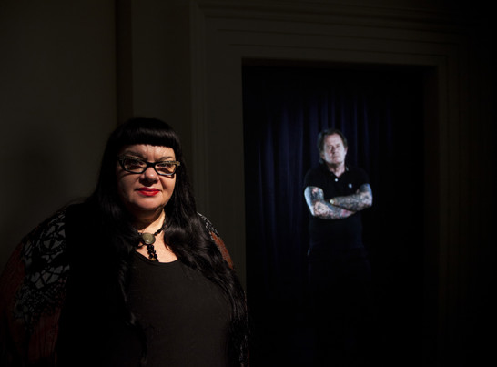 Woman who is in shadow standing in front of a screen displaying a man with tattoos on his arms