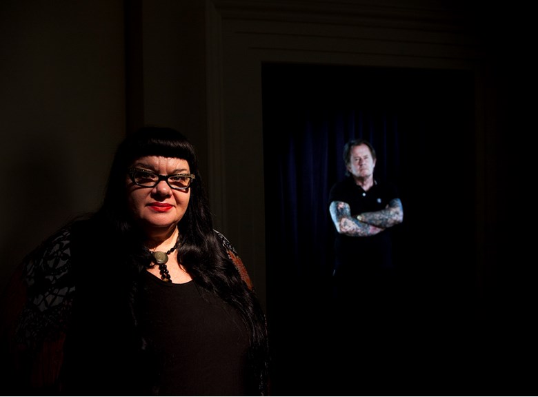 Woman who is in shadow standing in front of a screen displaying a man with tattoos on his arms