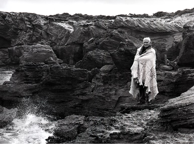 A man stands proudly on a cliff overlooking a wild ocean wrapped in a possum skin cloak.
