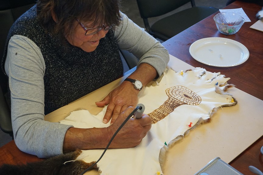 A woman sketches an eel basket onto a possum skin with a wire-nib burner.