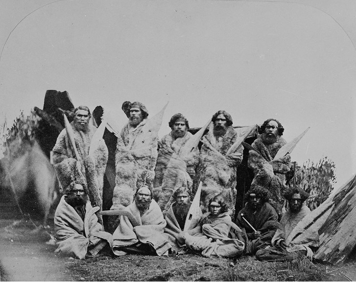 A group of men wrapped in possum skin cloaks and blankets. 