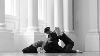Three performers on the floor in an intertwined contemporary dance pose beneath tall columns.