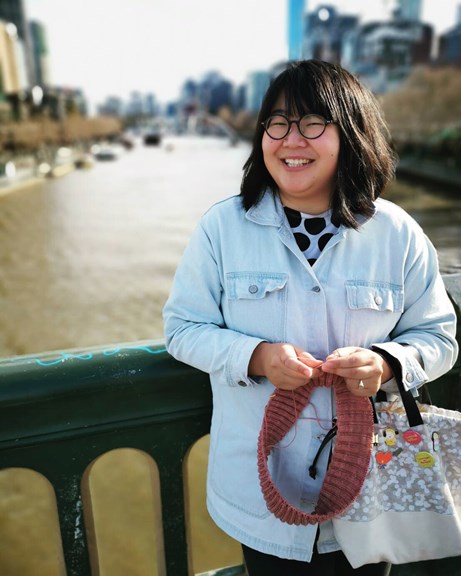 Sophia Cai, wearing a blue shirt, leans against a bridge railing, overlooking a river.