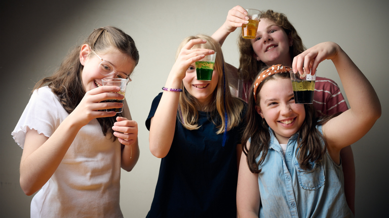 Four young people smile while conducting an experiment with different coloured liquids in plastic cups