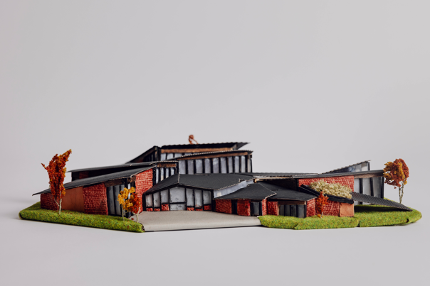 Architectural model of a building with red brick walls and black shopping roofs red brick walls, black sloped roofs, surrounded by trees with orange foliage.