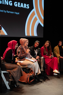 A group of Muslim women panellists, dressed mainly in blacks, reds and yellows, and holding mics, are spotlighted on a stage as they speak.