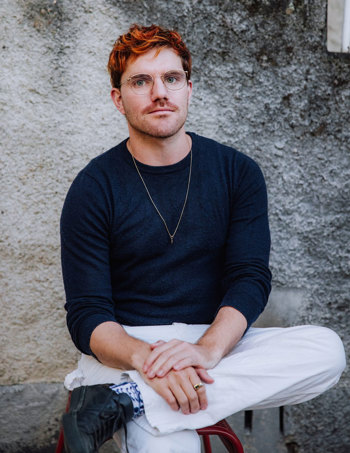 Brendan is sitting in a relaxed pose with the legs crossed and both hands resting on the leg. Brendan wears a dark navy long sleeve top and white pants.