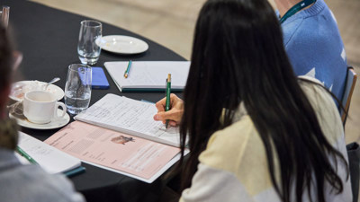 Pictured from the back of a womans head with long brown hair, seated at a table making notes. 