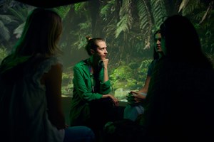 Women listen in a group of people sitting on stools. A large image of a forest covers the wall in the background.