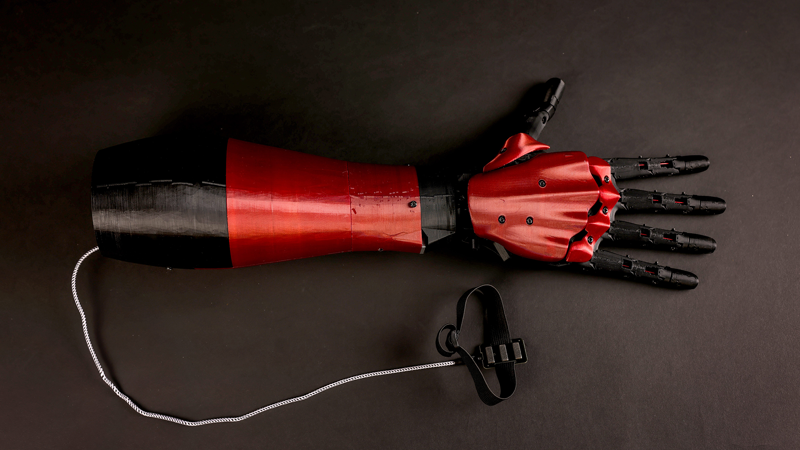 3D printed red and black prosthetic arm. A cable extends from the base of the arm, connecting to a sensor. 