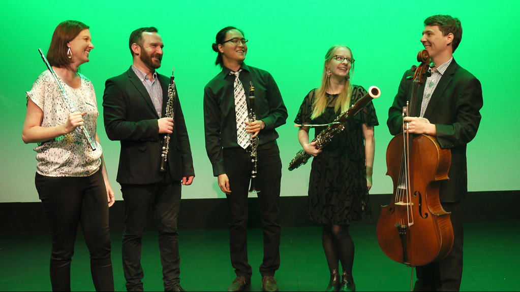 Five Musicians standing in front of a green wall each holding an instrument, including the flute, clarinet, oboe, bassoon and cello