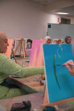 A classroom of Muslim women sit with mini easels and canvases in front of them, painting abstract blue and pink portraits. 