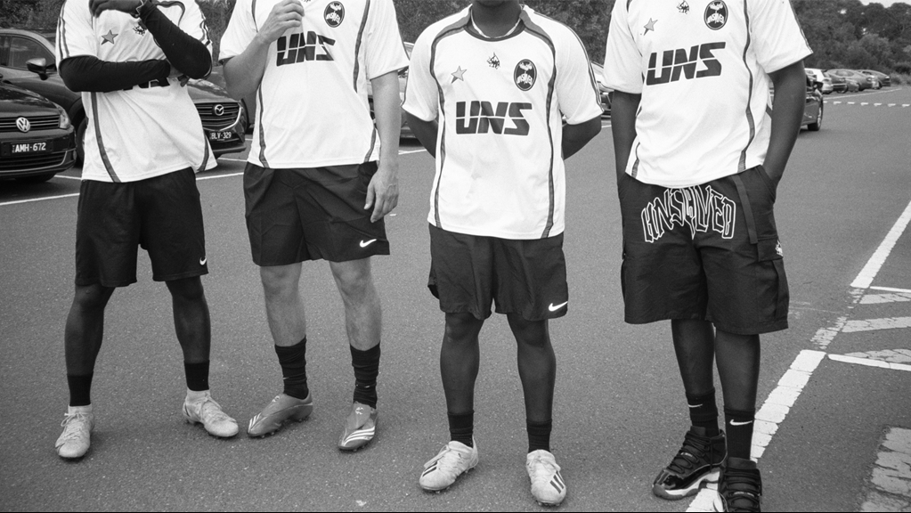 A black-and-white photograph of four young men standing in a parking lot, wearing matching white soccer jerseys with "UNS" printed on the front. Their faces are cropped out, focusing on their uniforms, shorts, and footwear, which include a mix of soccer cleats and sneakers. 