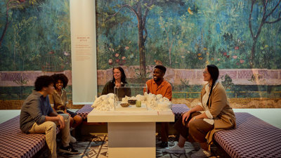 Group of visitors seated around a table exhibit, with a painted Roman garden mural filling the background. 
