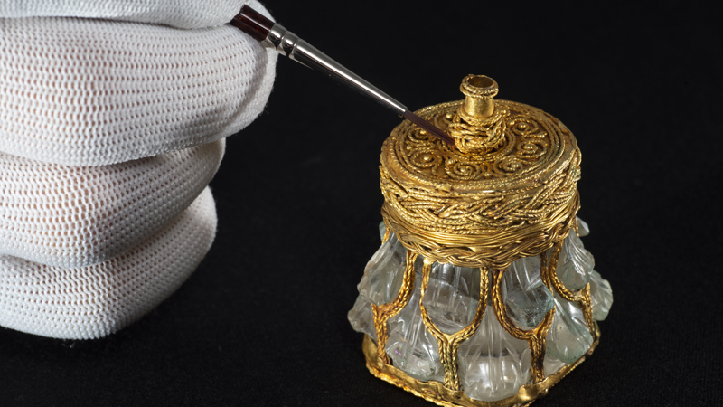 A hand uses a brush on the gold wire wrap makings of a rock crystal jar from the Galloway Hoard.