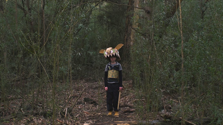 Film still showing a little boy wearing a homemade monster costume with a large toothy head and decorated chest piece, standing in the Australian bush and holding a toy sword.
