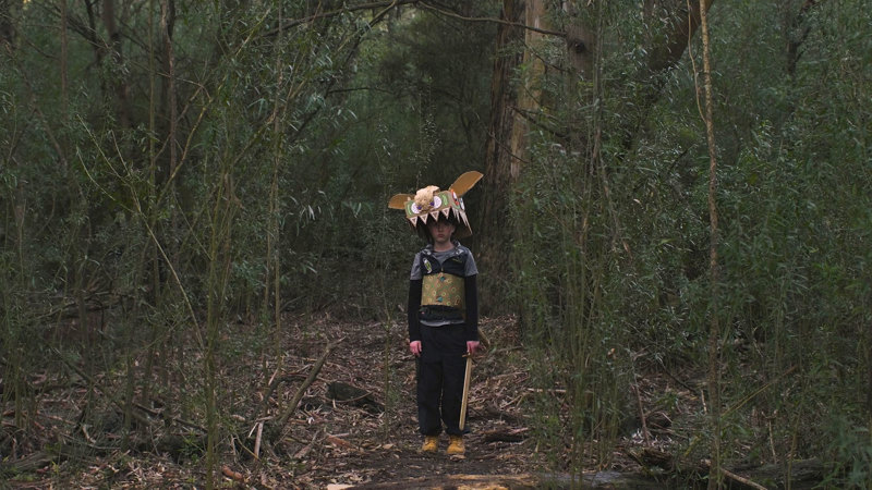Film still showing a little boy wearing a homemade monster costume with a large toothy head and decorated chest piece, standing in the Australian bush and holding a toy sword.