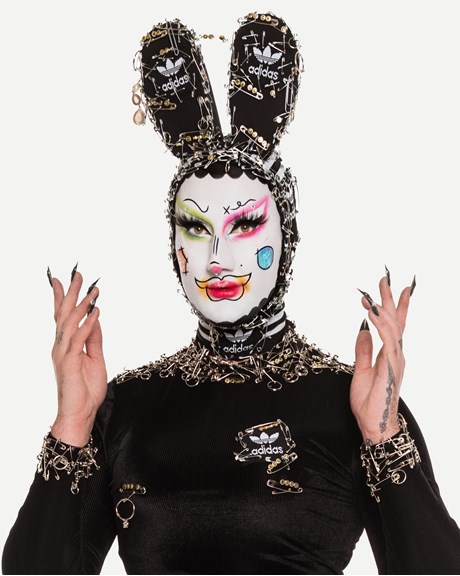 Simple the Drag Qween wearing black bejewelled bunny ears and vivid white makeup with colourful highlights. 