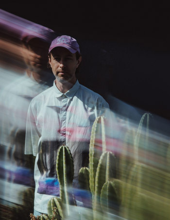 Ryan Powderly stands behind some cacti wearing a blue button up shirt and pink cap. The image is stylistically blurry and psychodelic.