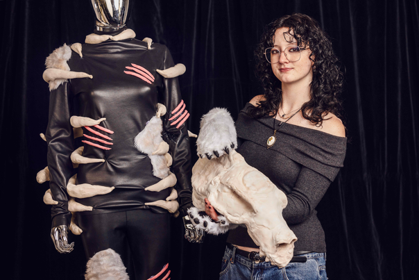Woman with curly hair and glasses, holding a large sculpted skull and standing next to a mannequin wearing a black costume with claw-like details.