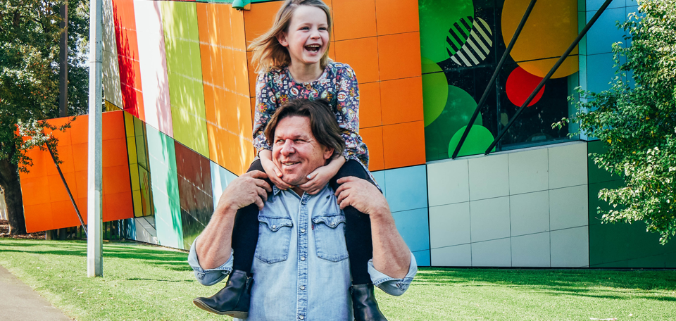 A man with a child on his shoulders walks along a path with the colourful walls to the Children's gallery in the background.